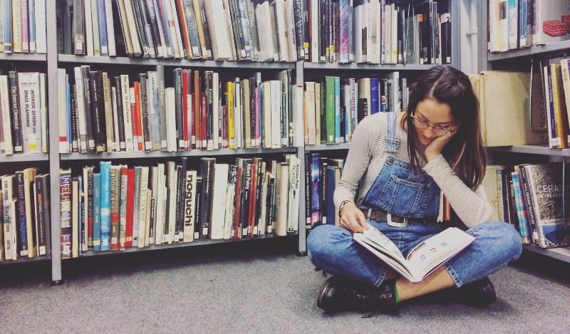 A college student reading a book on the floor in the library