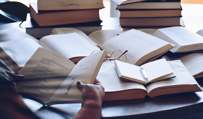 Woman is reading a book, taking notes in a notebook, multiple open books on a table with reading glasses on top ?