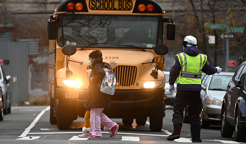 A school crossing guard holds a bus as a woman and child cross the street towards Public school 7 Louis F. Simeone in the Queens borough of New York, NY, December 8, 2020.